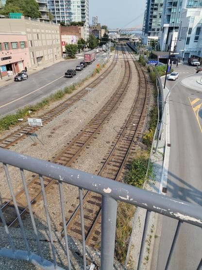 A photo of three slightly curved railway tracks between streets and dense buildings. In the distance there is a level crossing. The railing of a pedestrian overpass can be seen in the foreground.